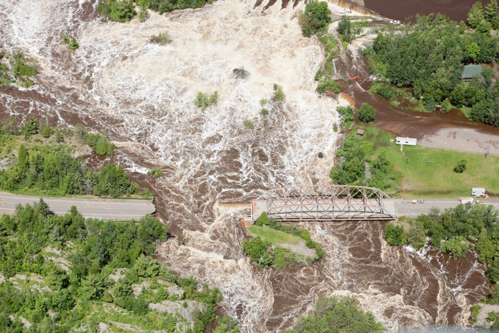 Aerial view of a two lane road washed away from storm floodwater with a truss bridge to the right. A stranded car is in the upper right Shot from the open window of a small airplane