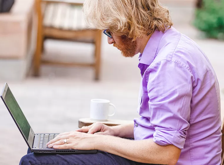 A person sitting outside, working on a laptop with cup of coffee. Exponent's engineering research advances next gen computer electronics.