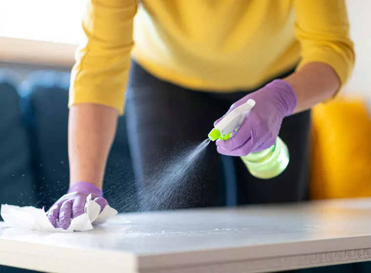 Hands in protective gloves holding spray bottle and disinfecting coffee table