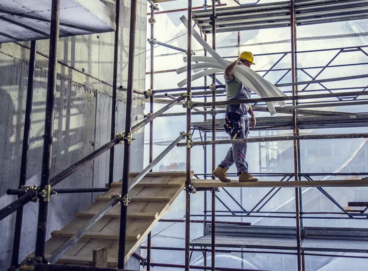 Worker balances PVC pipe while walking on scaffolding at a construction site. Exponent investigates construction accidents.