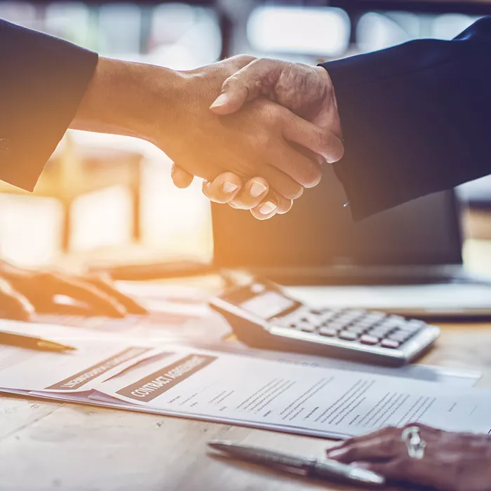 Two people shake hands over a table with contract agreement paperwork laying on top  