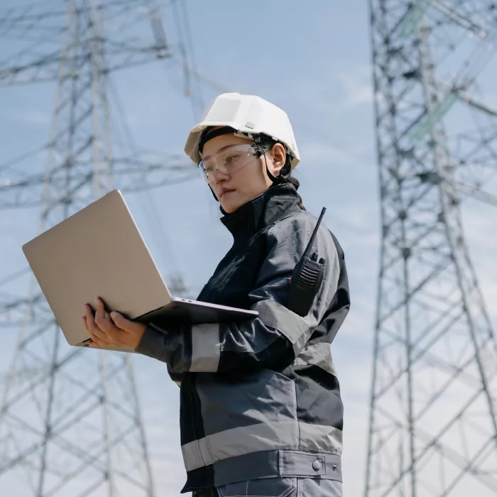 A utilities professional wearing goggles and helmet, staring at a laptop with 2 electrical towers in background