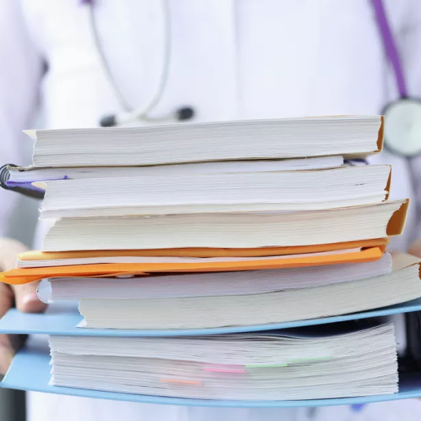 A healthcare worker holds a large stack of paperwork and folders while wearing a stethoscope around their neck