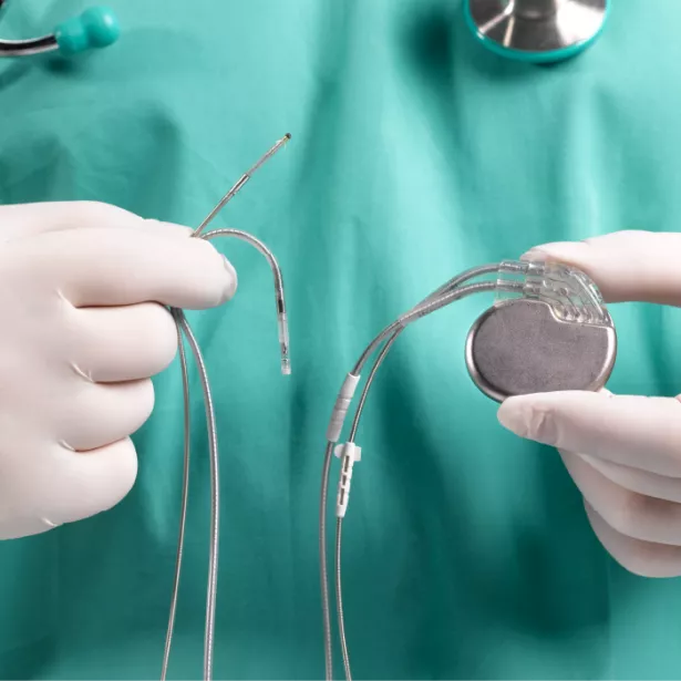 Gloved, medical professional in green scrubs with stethoscope around neck (shown from neck to mid torso) holding an implanted medical device