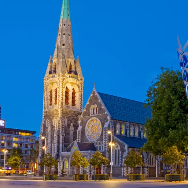 Old ornamental church lit up at night between a modern high-rise building and modern art sculpture of an inverted cone, after light snowfall