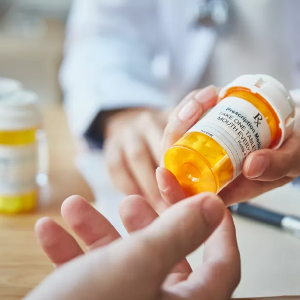 Medical professional handing a prescription bottle to a patient over a table with prescription bottles and pens in the background