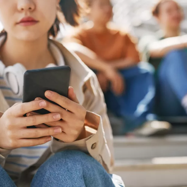 Young woman with headphones around her neck looking at her cell phone screen while sitting in the bleachers with a row of young people sitting in the background