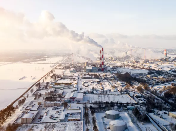 Aerial view of snow-covered industrial section of city with red and white striped smokestacks blowing smoke across hazy, dim-lit sky