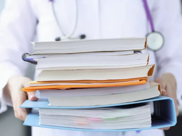 A healthcare worker holds a large stack of paperwork and folders while wearing a stethoscope around their neck