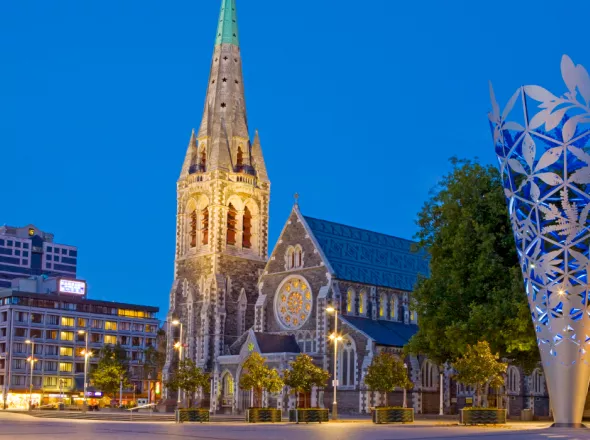 Old ornamental church lit up at night between a modern high-rise building and modern art sculpture of an inverted cone, after light snowfall