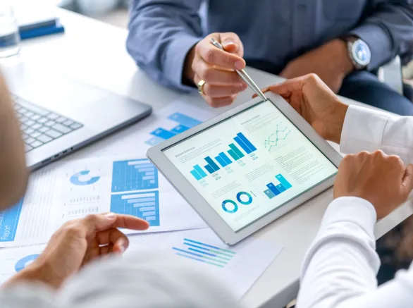 Close up of three people looking at financial data with graphs and charts. All their hands can be seen and one person is pointing with a pen. There is paperwork on the desk showing more finance information