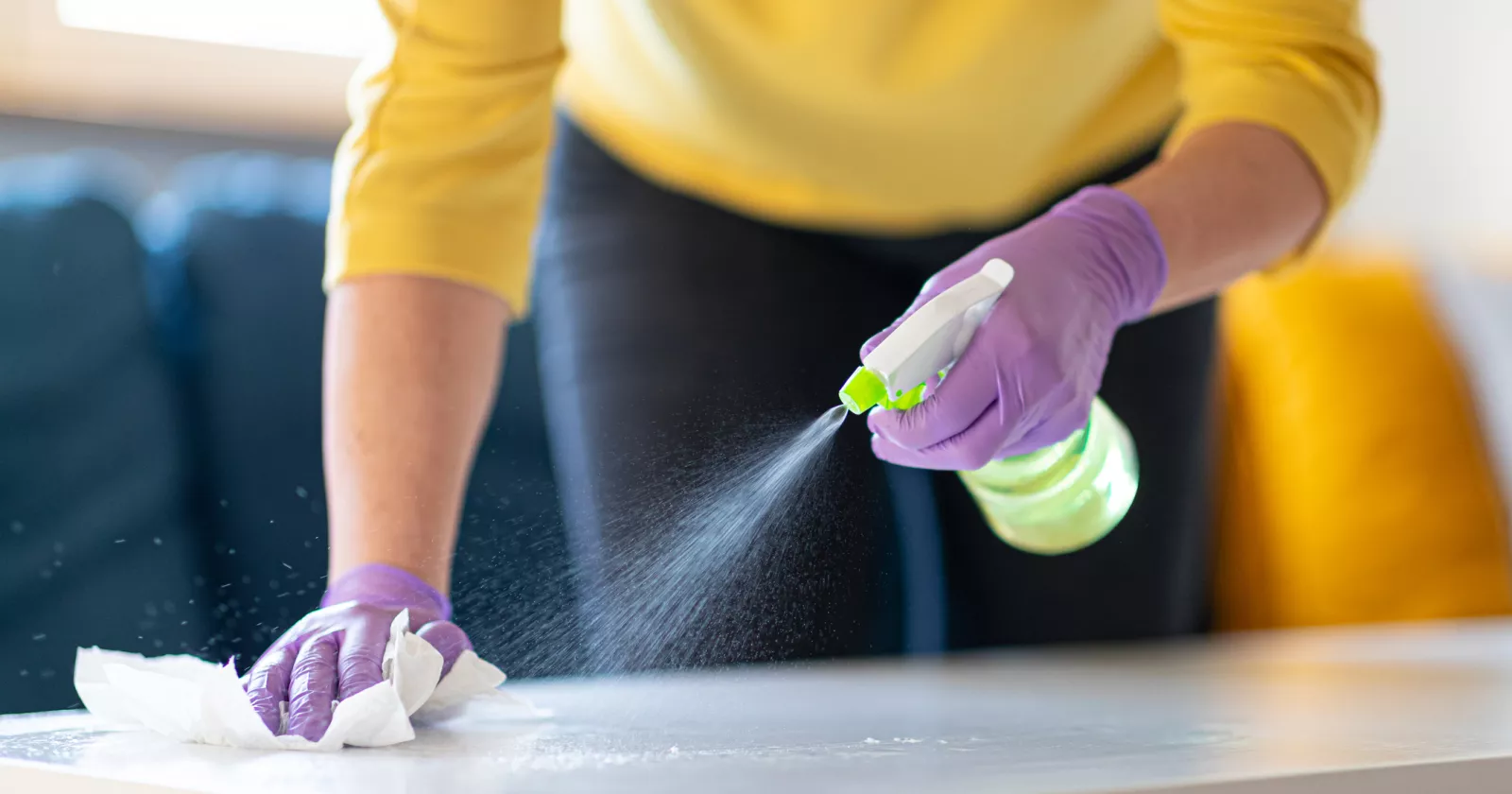 Hands in protective gloves holding spray bottle and disinfecting coffee table 