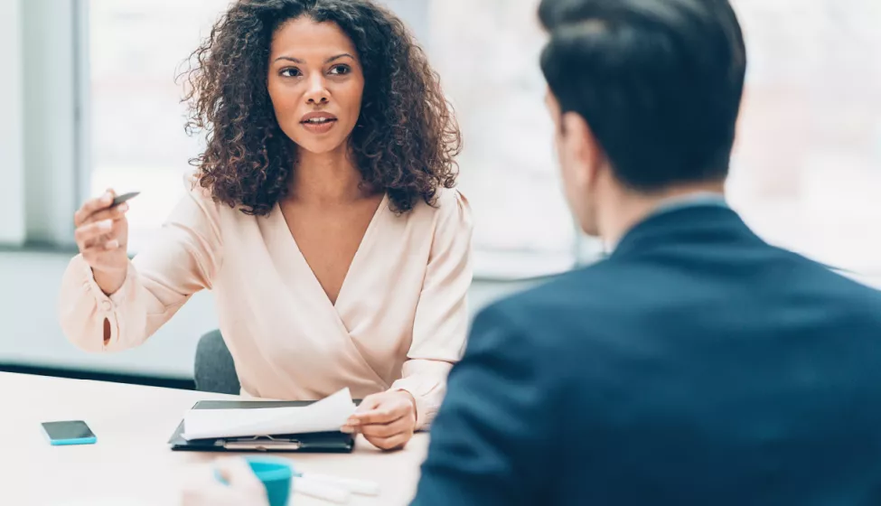 Two business people sitting at a table across from one another engaged in conversation--one is holding a pen and papers