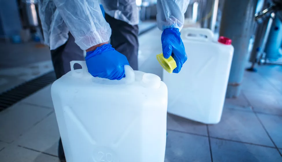 Worker in transparent jacket and blue gloves removing yellow cap from large plastic rectangle container inside a plant