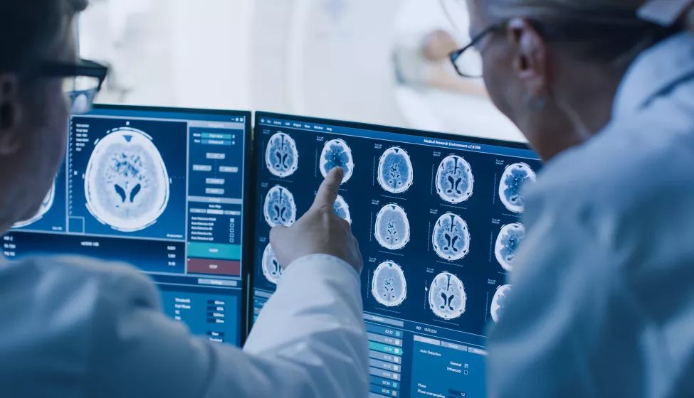 In Control Room Doctor and Radiologist Discuss Diagnosis while Watching Procedure and Monitors Showing Brain Scans Results, In the Background Patient Undergoes MRI or CT Scan Procedure