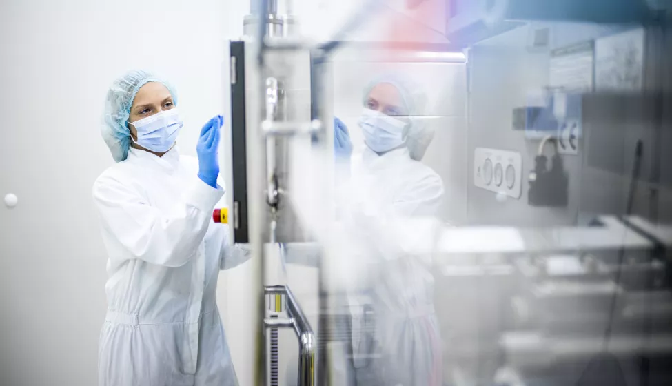 A woman wearing proper safety clothes seen taping on the screen of a big complex machine in pharmaceutical industry while her reflection is visible on the machine.