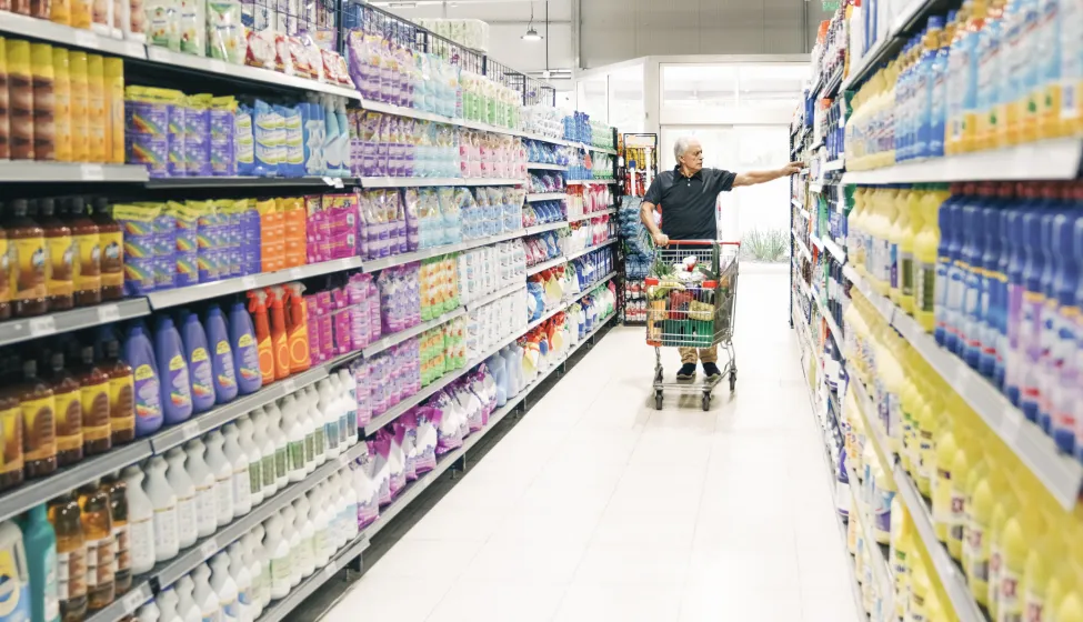 Senior man with shopping cart in supermarket aisle. Mature male customer looking at groceries in store.