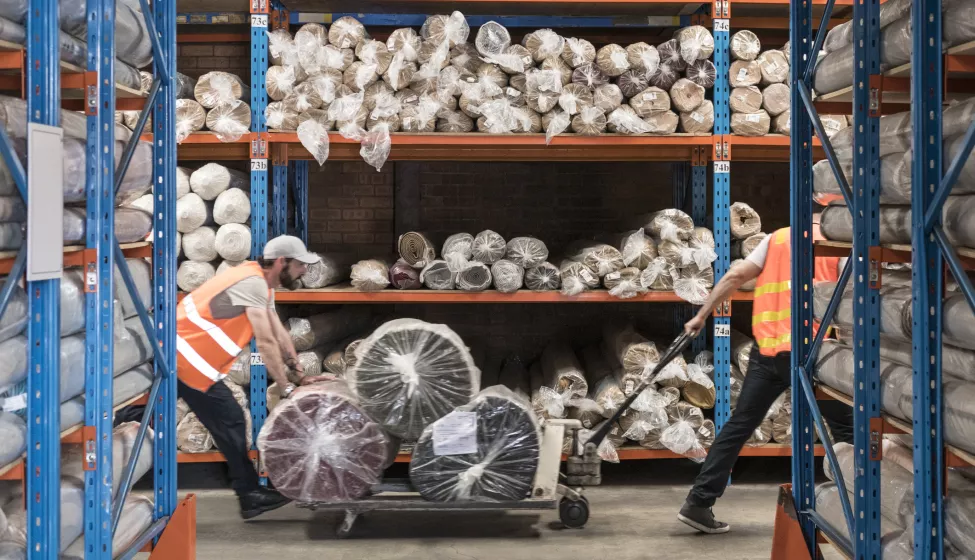 Two workers in a warehouse pushing a cart of plastic packages, surrounded by shelves of more plastic packages