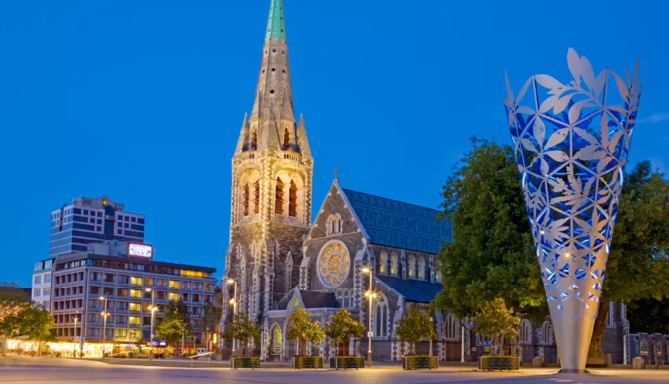 Old ornamental church lit up at night between a modern high-rise building and modern art sculpture of an inverted cone, after light snowfall