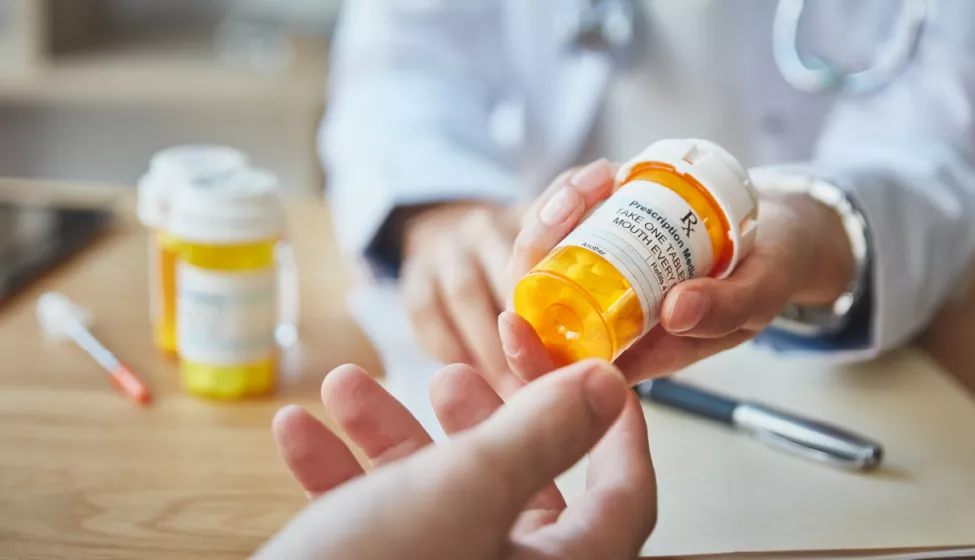 Medical professional handing a prescription bottle to a patient over a table with prescription bottles and pens in the background