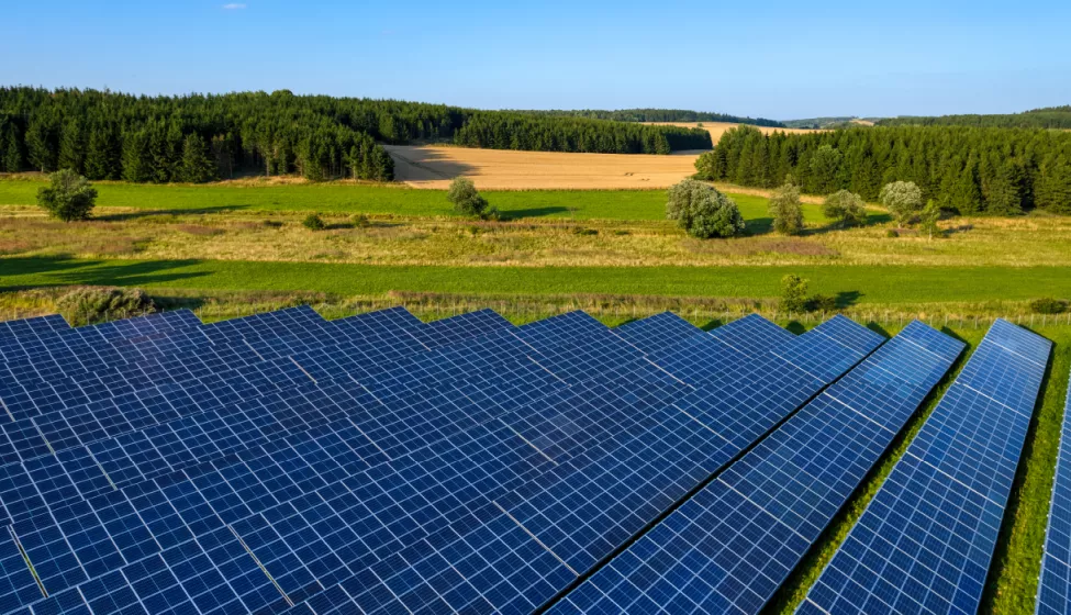 Solar panels in meadow with green grass and tree-covered hillside with building in the background